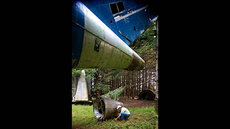 Campbell picks up small fragments of blue paint that fell when he recently pressure-washed the fuselage. Pressure-washing is a regular maintenance duty in wet and forested areas of the Pacific Northwest, though Campbell says the task goes quickly on the smooth-skinned aircraft. On the ground next to Campbell is a thrust-reverser from the rear engine, used for decelerating the plane after landing.